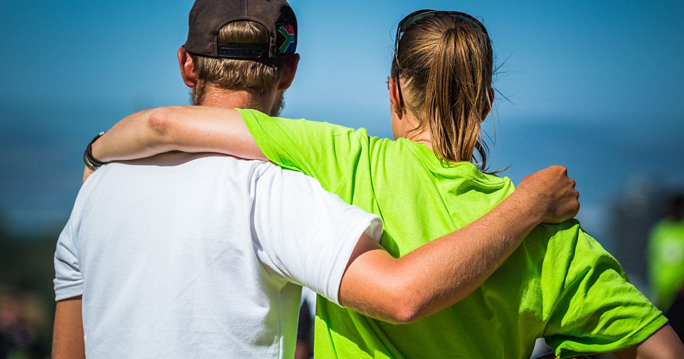 Two volunteer sports coaches have their arms around each other's shoulders, watching the exercise.