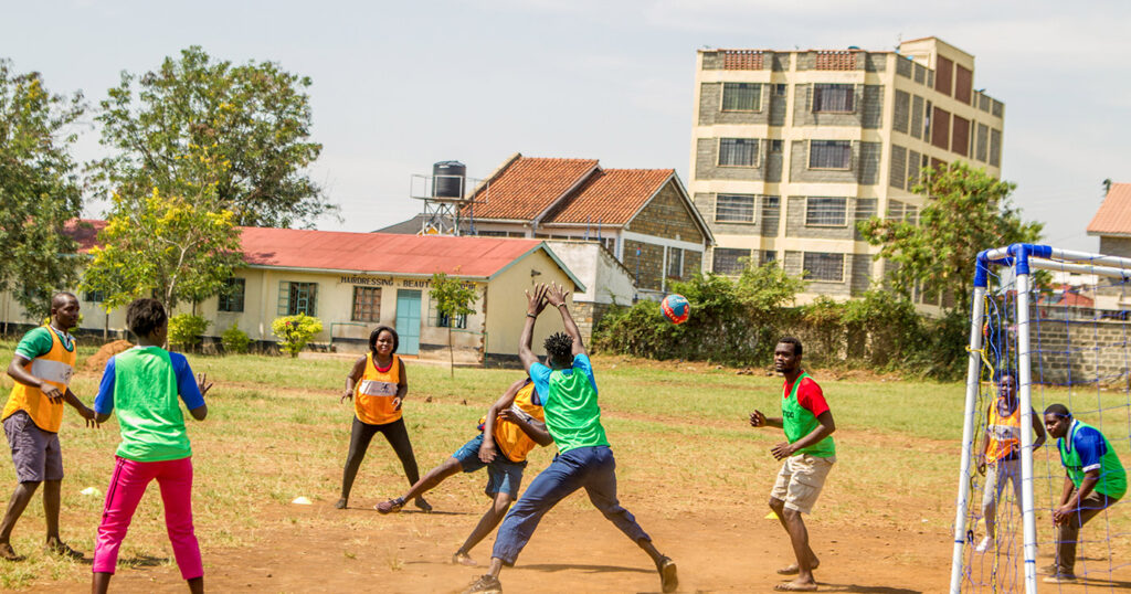 A mixed youth handball team is in the midst of an attack in front of the goal on a dry grass pitch in a residential area.