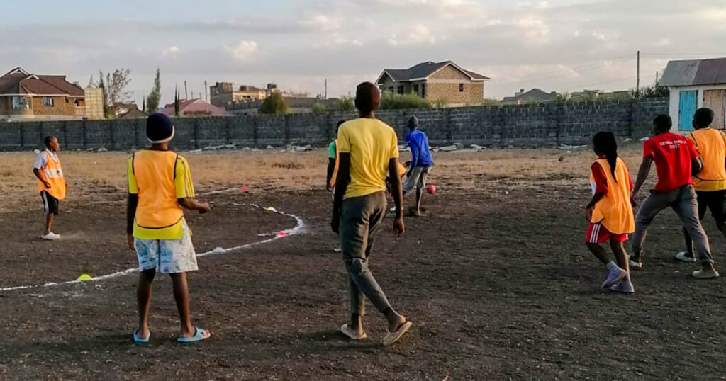 A group of children and teenagers in flip-flops are practising handball in a residential area on an open space with a bumpy and stony surface.