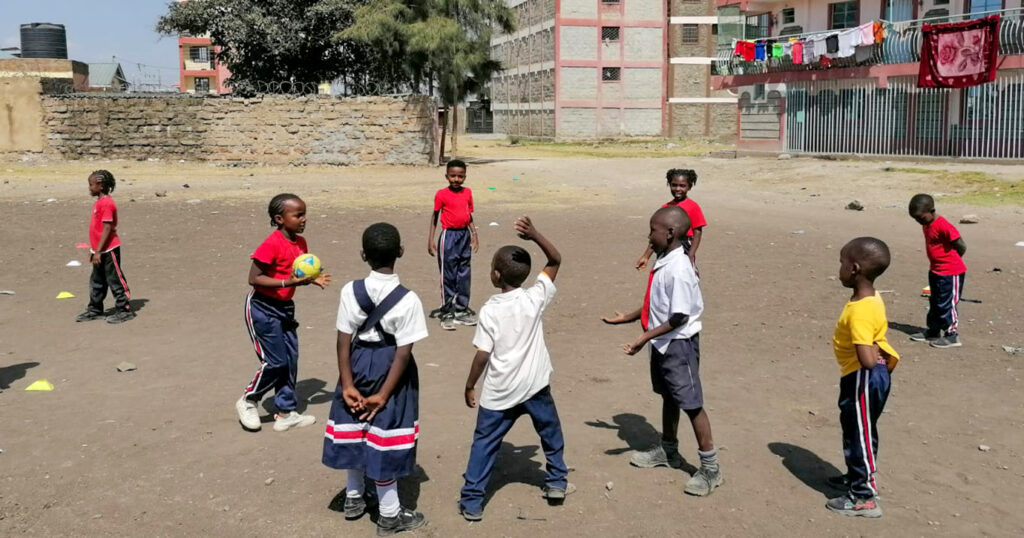 A group of boys and girls play handball together on a dusty court in front of an apartment building.