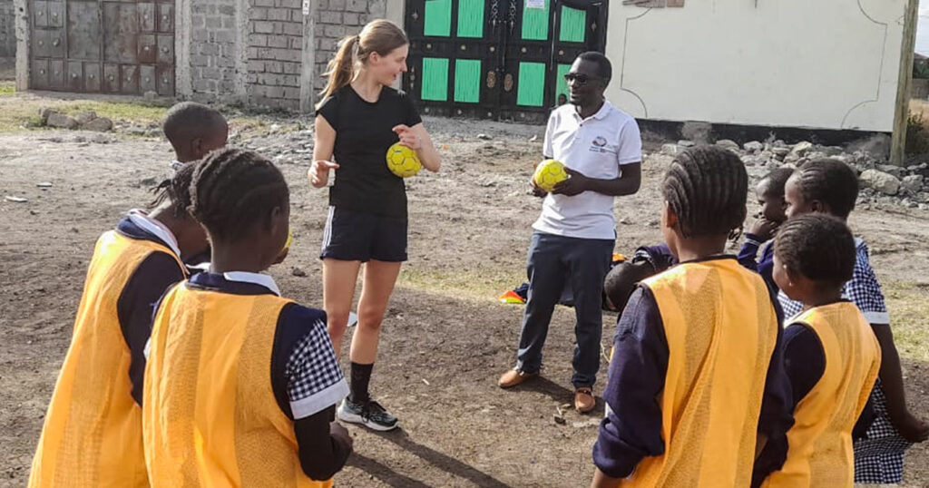 Eine ehrenamtliche Handballtrainerin unterrichtet einen lokalen Handballtrainer, umringt von Kindern aus ihrem Projekt.