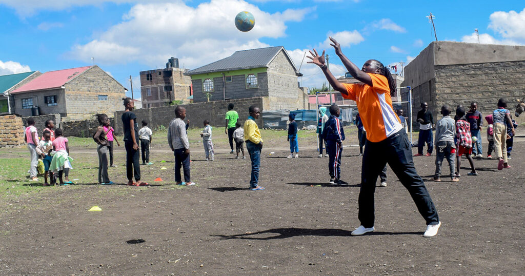 A player catches a handball while a large group of children stand in the background watching a handball coach give a lesson.