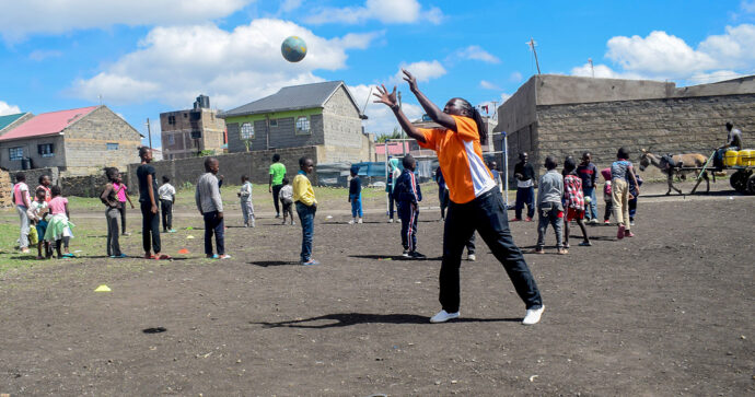 Eine Spielerin fängt einen Handball, während im Hintergrund eine große Gruppe von Kindern steht und einem Handballtrainer beim Unterrichten zusieht.