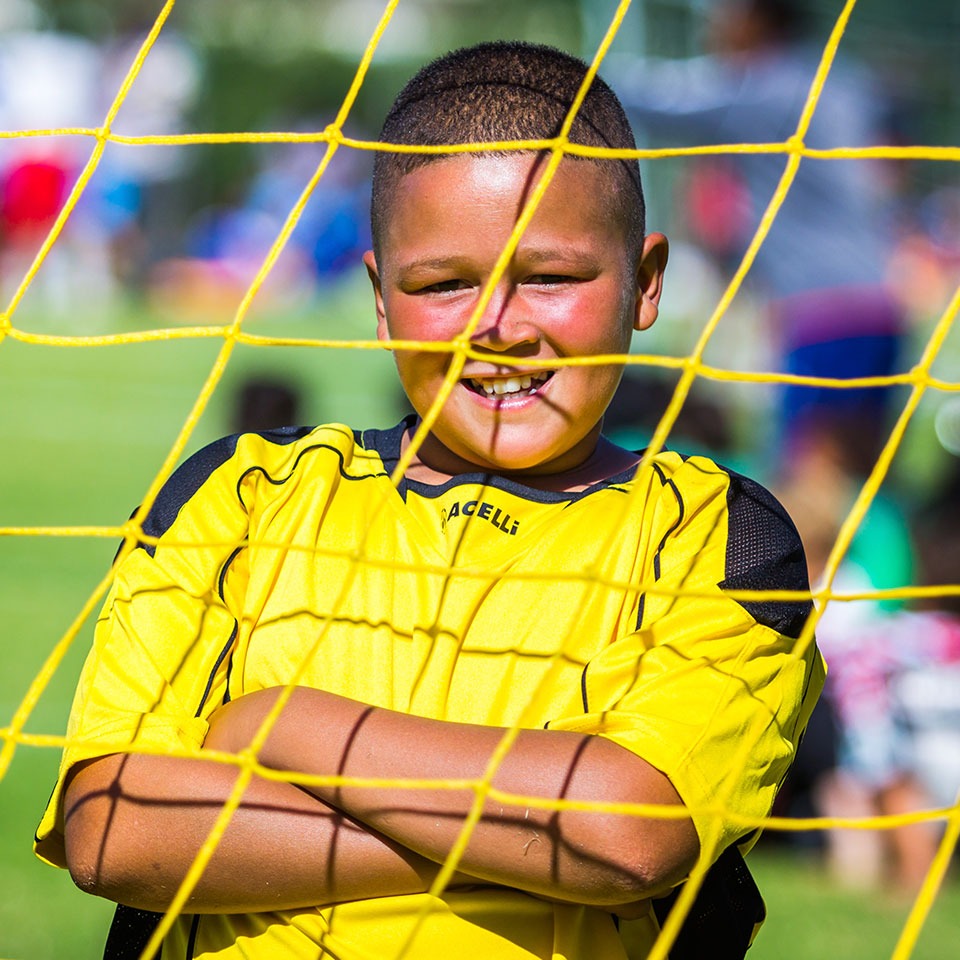 A boy in a sports shirt smiles and poses for the camera with his arms folded behind a goal net.