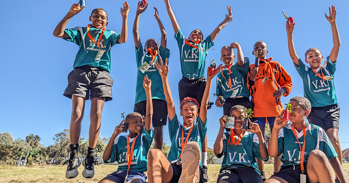 Children from a sports team leap into the air with their hands in the air, holding their trophies.