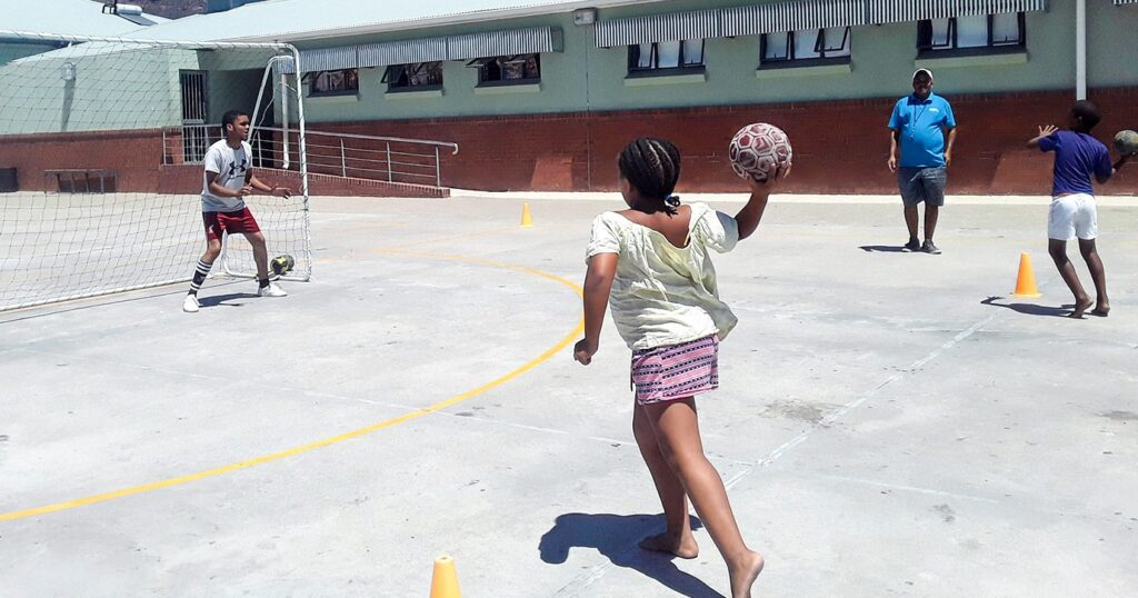 A girl and a boy simultaneously throw a handball at a goal with a goalkeeper for training purposes on a concrete school sports field, under the eyes of the sports project leader.