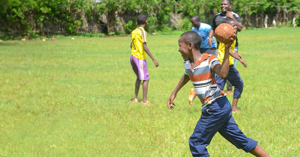 On a green lawn, a boy in street clothes throws a handball during sports training, while other boys continue to play with the project mentor in the background.