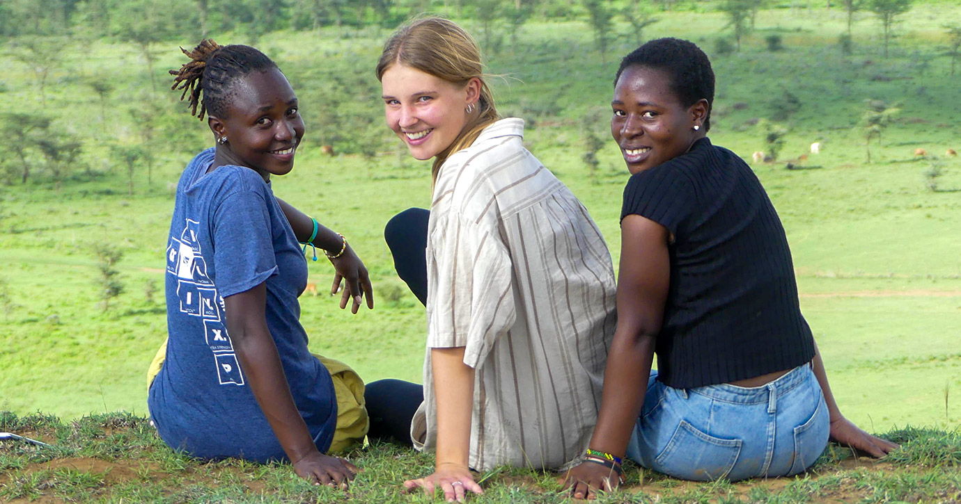 A German volunteer in Kenya poses with two local women in a green landscape.